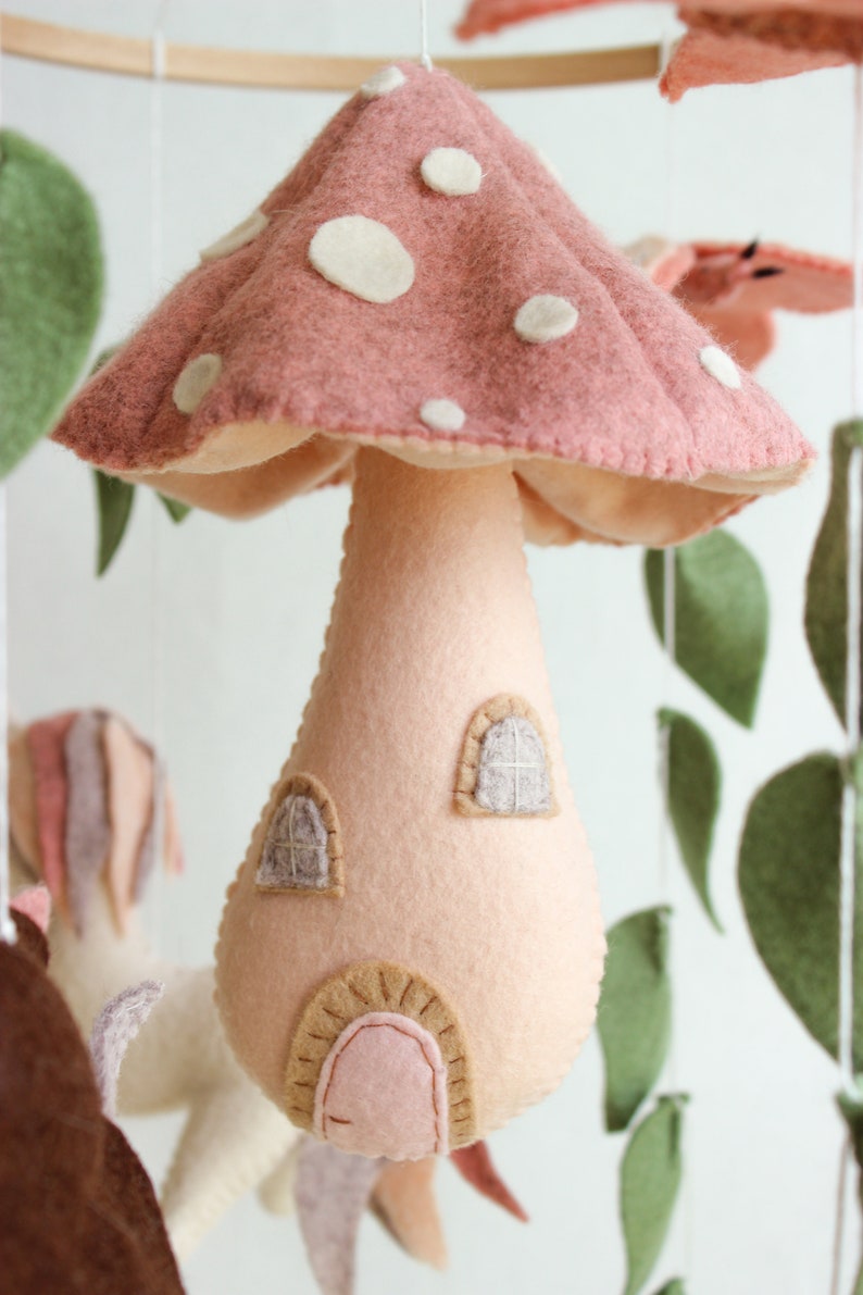 May include: A pink felt mushroom with white polka dots and a small house-shaped stem. The mushroom has two windows and a door.