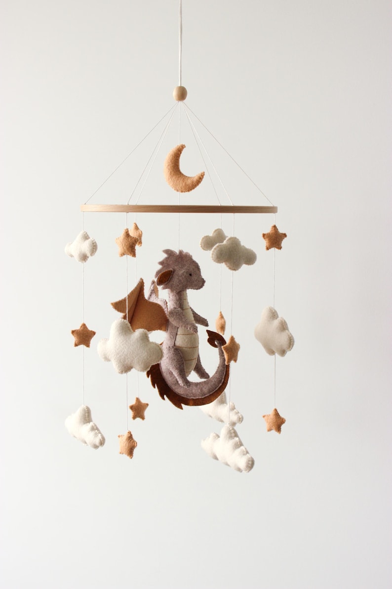 May include: A baby mobile featuring a felt dragon, clouds, stars, and a crescent moon. The dragon is lavender with brown wings and tail. The clouds and stars are white and tan, suspended from a wooden ring.