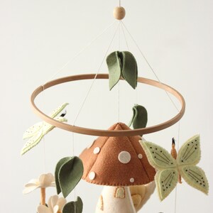 May include: A whimsical baby mobile featuring felted woodland creatures. The mobile includes a brown mushroom with white spots, green leaves, a butterfly, a frog, and a beetle, all suspended from a wooden hoop.