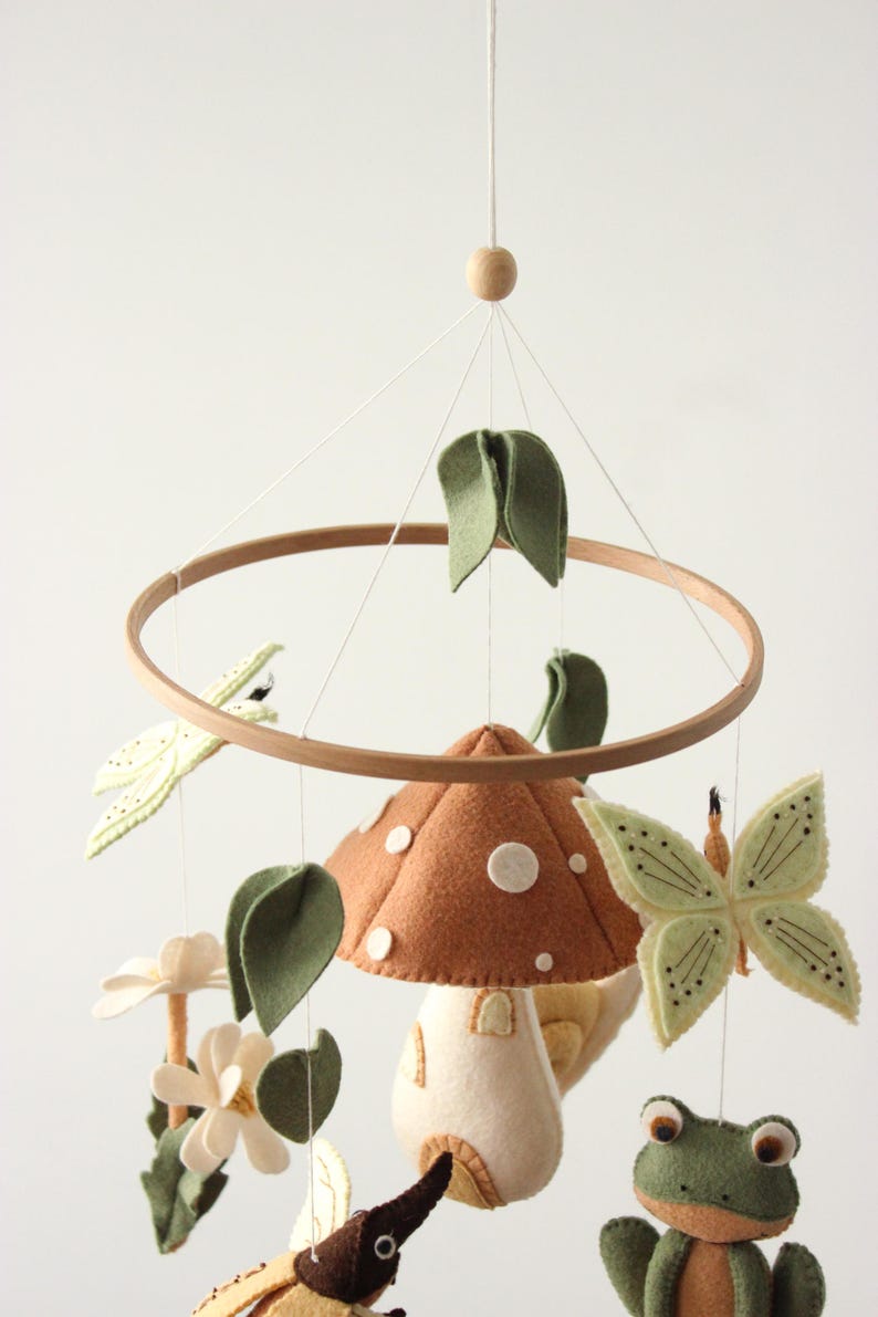 May include: A whimsical baby mobile featuring felted woodland creatures. The mobile includes a brown mushroom with white spots, green leaves, a butterfly, a frog, and a beetle, all suspended from a wooden hoop.