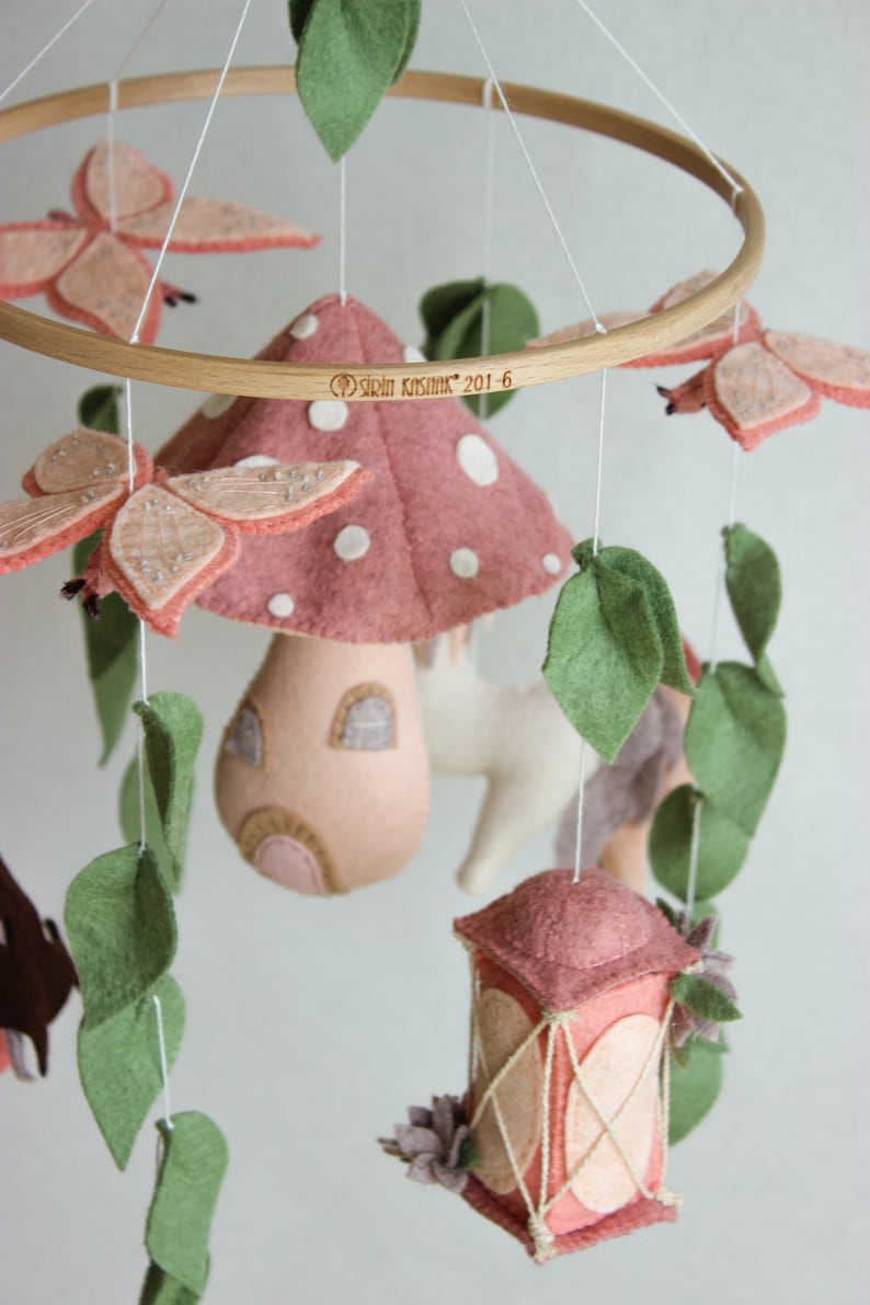 May include: A pink and white felt mushroom mobile with a small house, a lantern, butterflies, and leaves. The mobile is hanging from a wooden ring and has the text "Sril Kasimir 201-6" on it.