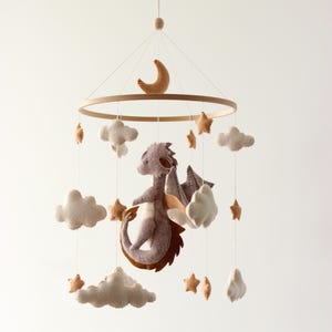May include: A baby mobile featuring a felt dragon, clouds, stars, and a crescent moon. The dragon is purple with brown accents and wings. The clouds and stars are beige. The mobile hangs from a wooden ring.