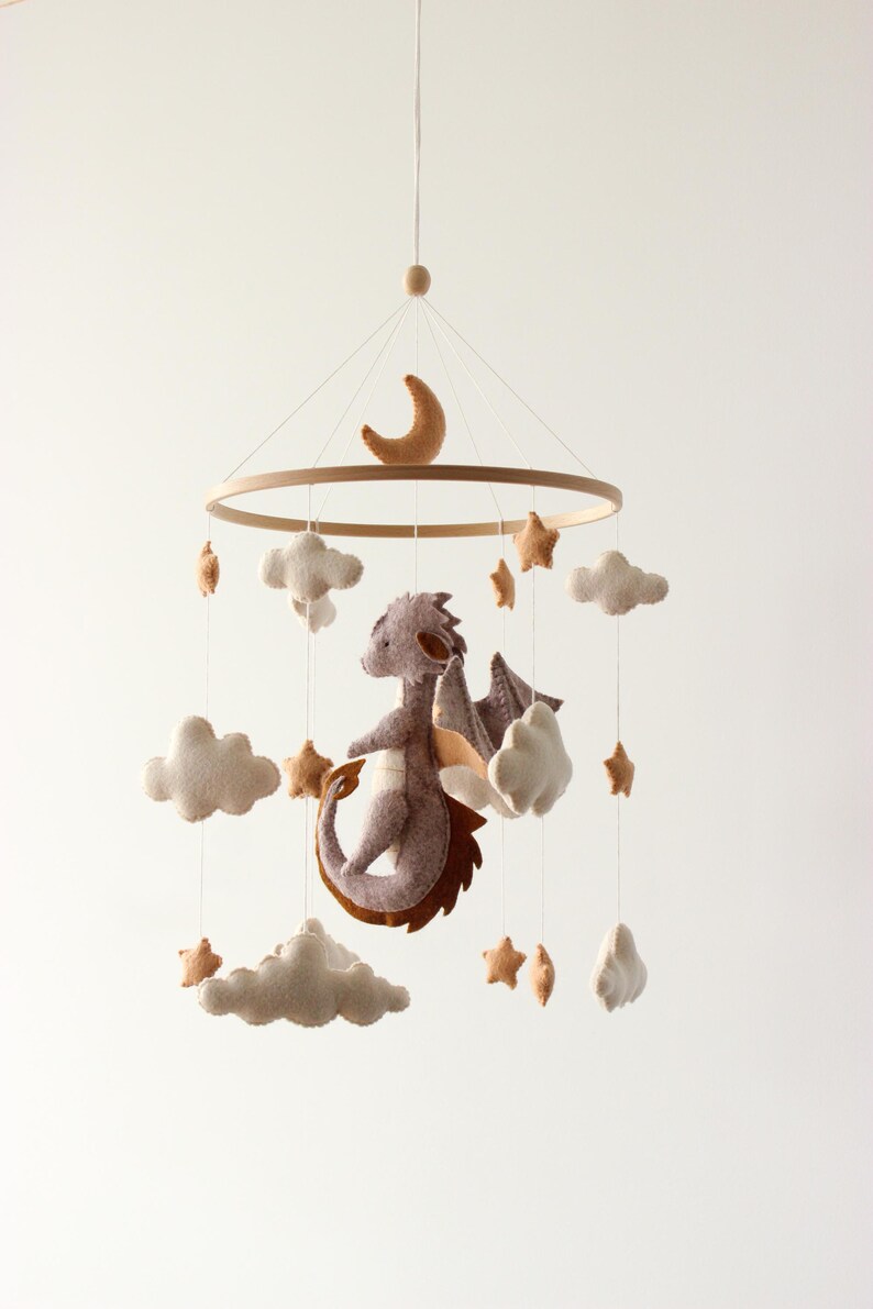 May include: A baby mobile featuring a felt dragon, clouds, stars, and a crescent moon. The dragon is purple with brown accents and wings. The clouds and stars are beige. The mobile hangs from a wooden ring.