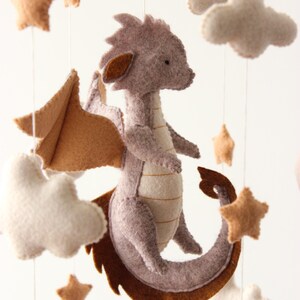 May include: A handmade baby mobile featuring a felt dragon in shades of gray and brown, with matching stars and fluffy white clouds. The dragon has large wings and a long tail. The mobile is suspended from a wooden frame.