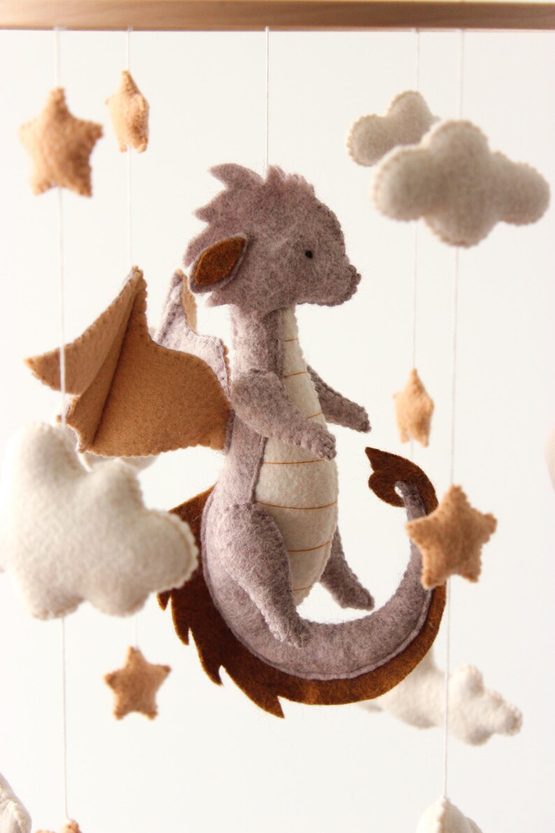 May include: A handmade baby mobile featuring a felt dragon in shades of gray and brown, with matching stars and fluffy white clouds. The dragon has large wings and a long tail. The mobile is suspended from a wooden frame.