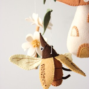 May include: A handmade felt mobile featuring a brown and tan beetle with cream-colored wings, a mushroom house, flowers, and leaves. The mobile is suspended by white strings against a white background. The beetle has stitched details.
