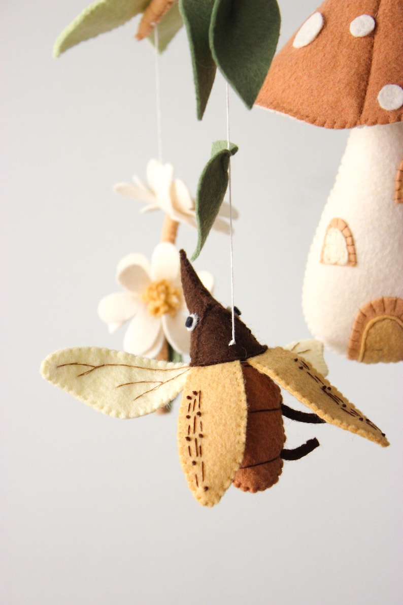 May include: A handmade felt mobile featuring a brown and tan beetle with cream-colored wings, a mushroom house, flowers, and leaves. The mobile is suspended by white strings against a white background. The beetle has stitched details.