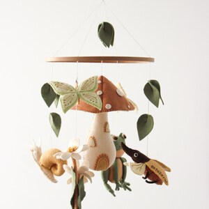 May include: A whimsical baby mobile featuring felted woodland creatures. The mobile includes a mushroom, butterfly, snail, frog, and beetle, along with green leaves. The creatures are suspended from a wooden ring, creating a charming nursery decoration.