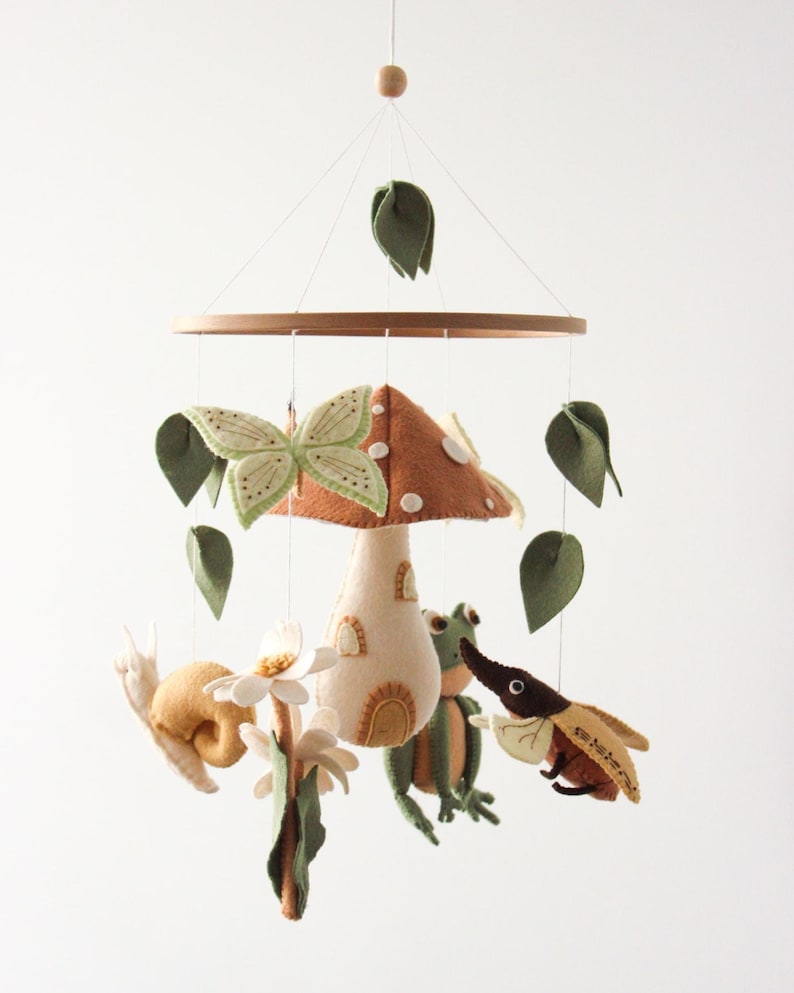 May include: A whimsical baby mobile featuring felted woodland creatures. The mobile includes a mushroom, butterfly, snail, frog, and beetle, along with green leaves. The creatures are suspended from a wooden ring, creating a charming nursery decoration.