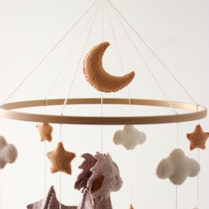 May include: A baby mobile featuring a wooden ring with felt shapes. The mobile includes a crescent moon, stars, clouds, and a dragon in shades of brown, tan, and white. The mobile hangs from a white string.