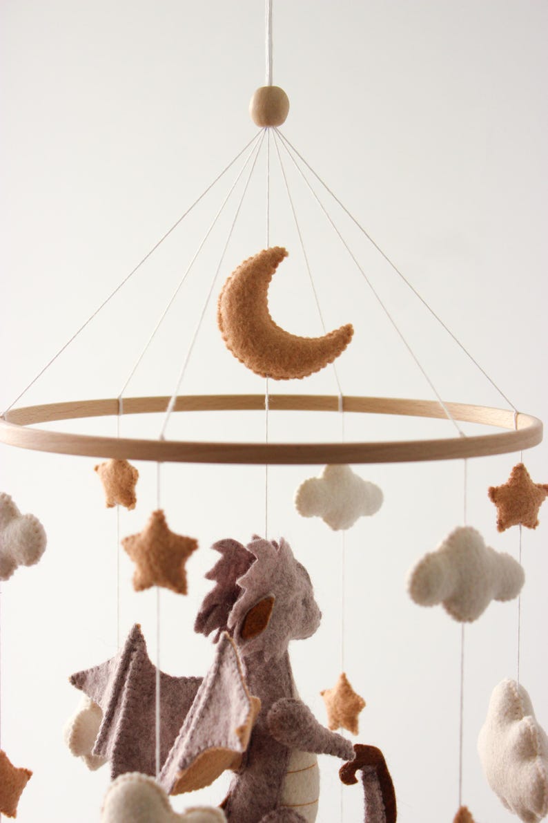 May include: A baby mobile featuring a wooden ring with felt shapes. The mobile includes a crescent moon, stars, clouds, and a dragon in shades of brown, tan, and white. The mobile hangs from a white string.