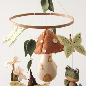 May include: A whimsical felt baby mobile featuring a wooden hoop, a mushroom, butterflies, a frog, and leaves. The mushroom is brown with white spots, and the butterflies and leaves are green. The mobile is suspended by white strings.