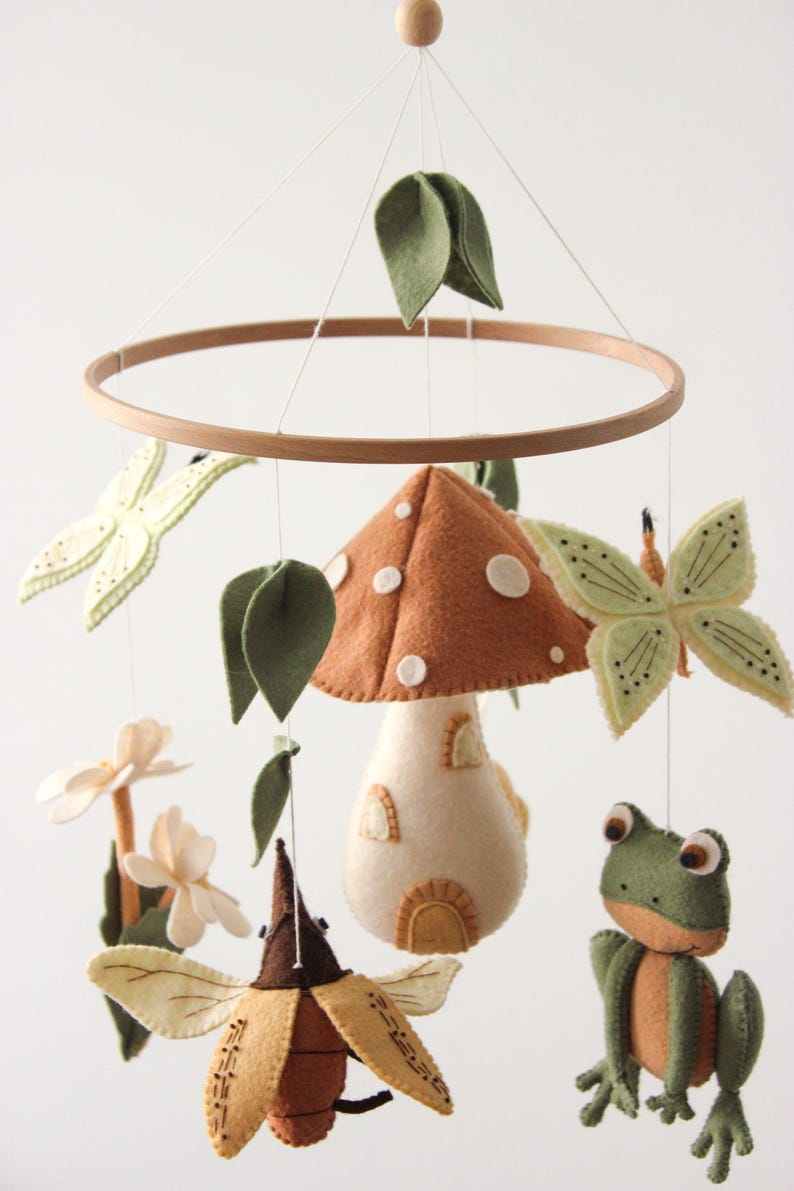 May include: A whimsical felt baby mobile featuring a wooden hoop, a mushroom, butterflies, a frog, and leaves. The mushroom is brown with white spots, and the butterflies and leaves are green. The mobile is suspended by white strings.
