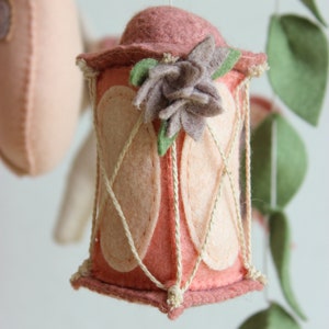 May include: A pink felt lantern with a white rope design and a brown felt flower on top. The lantern is hanging from a white string.