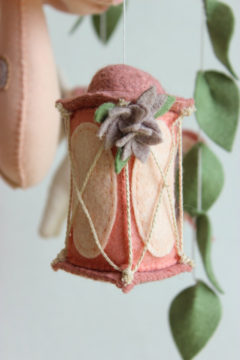 May include: A pink felt lantern with a white rope design and a brown felt flower on top. The lantern is hanging from a white string.