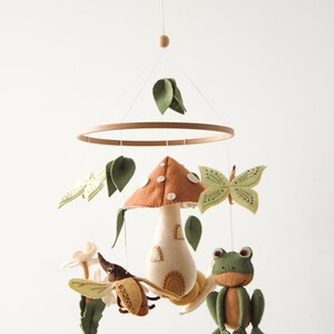 May include: A whimsical baby mobile featuring a mushroom, frog, butterfly, and beetle. The mobile has a wooden ring and is suspended by a string. The mushroom has a brown cap and a cream-colored stem. The frog is green and orange.