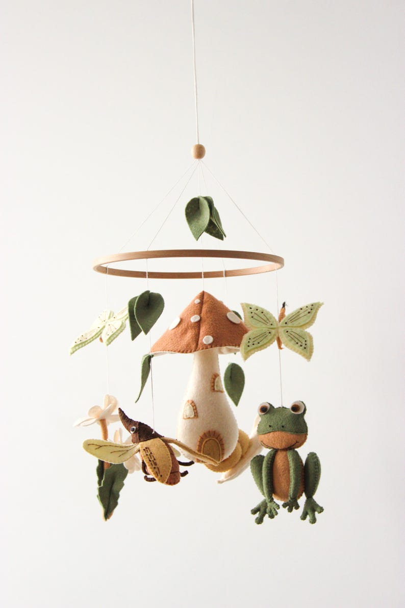 May include: A whimsical baby mobile featuring a mushroom, frog, butterfly, and beetle. The mobile has a wooden ring and is suspended by a string. The mushroom has a brown cap and a cream-colored stem. The frog is green and orange.