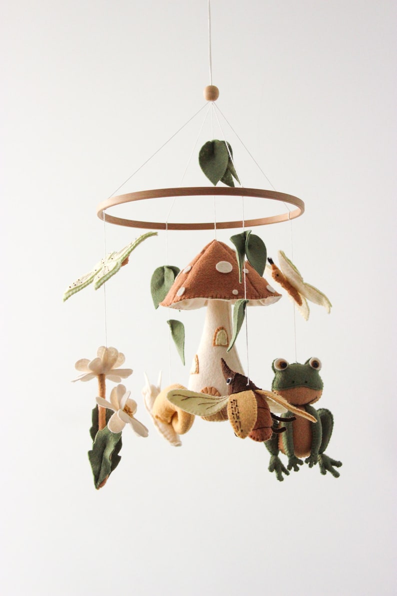 May include: A whimsical felt mobile featuring a mushroom house, a frog, bees, butterflies, and flowers. The mobile is suspended from a wooden ring and includes green leaves and delicate white flowers. The color palette includes shades of brown, green, and cream.