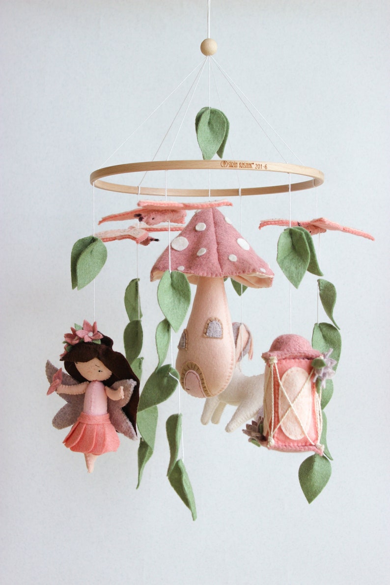 May include: A pink and green felt baby mobile with a mushroom house, a fairy, a unicorn, a drum, and leaves. The mobile is hanging from a wooden ring.