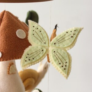 May include: A handmade baby mobile featuring felted woodland creatures. The mobile includes a mushroom, a butterfly with light green wings, a snail, and a frog. The mobile is suspended from a wooden hoop.