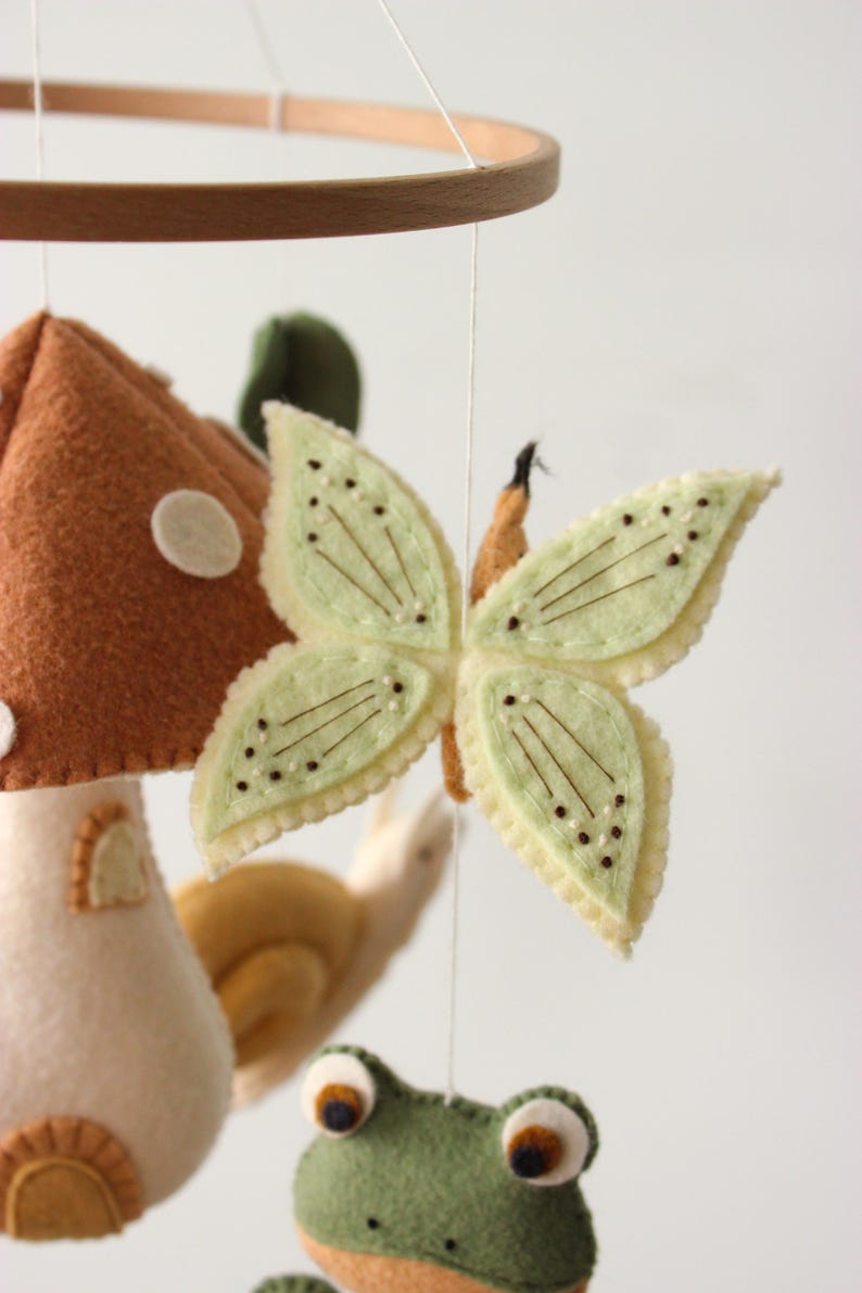 May include: A handmade baby mobile featuring felted woodland creatures. The mobile includes a mushroom, a butterfly with light green wings, a snail, and a frog. The mobile is suspended from a wooden hoop.