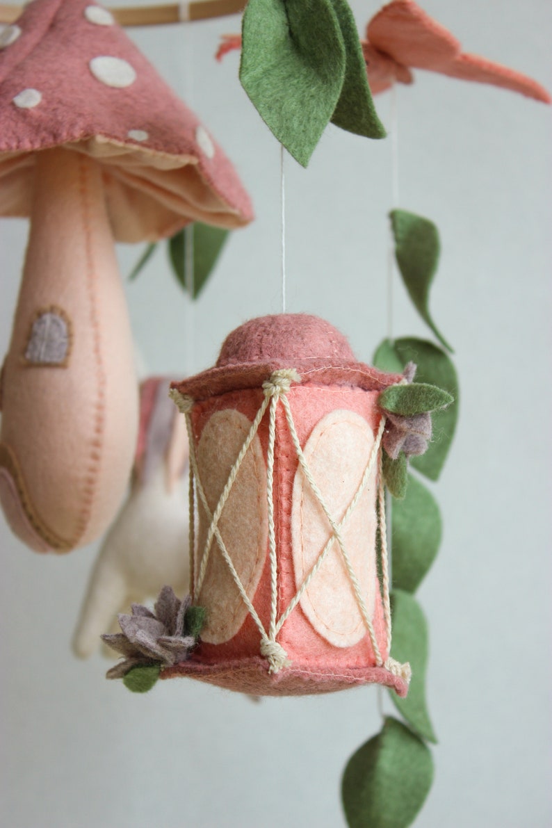 May include: A pink felt lantern with a white rope design hangs from a mobile. The lantern has a pink flower on the side.