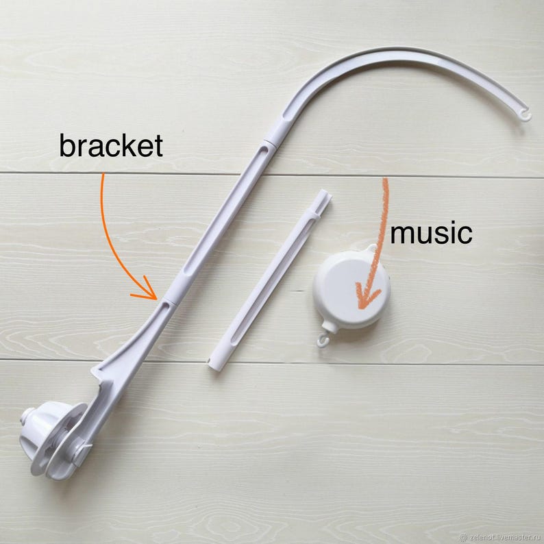 May include: A disassembled white baby mobile arm and music box, resting on a light wood surface. The mobile arm is curved and has a bracket. The music box is round and white. The word "music" is written on the image.