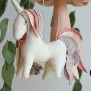 May include: A white felt unicorn with a pink and brown mane and tail hangs from a mobile. The unicorn is suspended from a white string. The mobile also features a pink felt mushroom with white spots and green felt leaves.