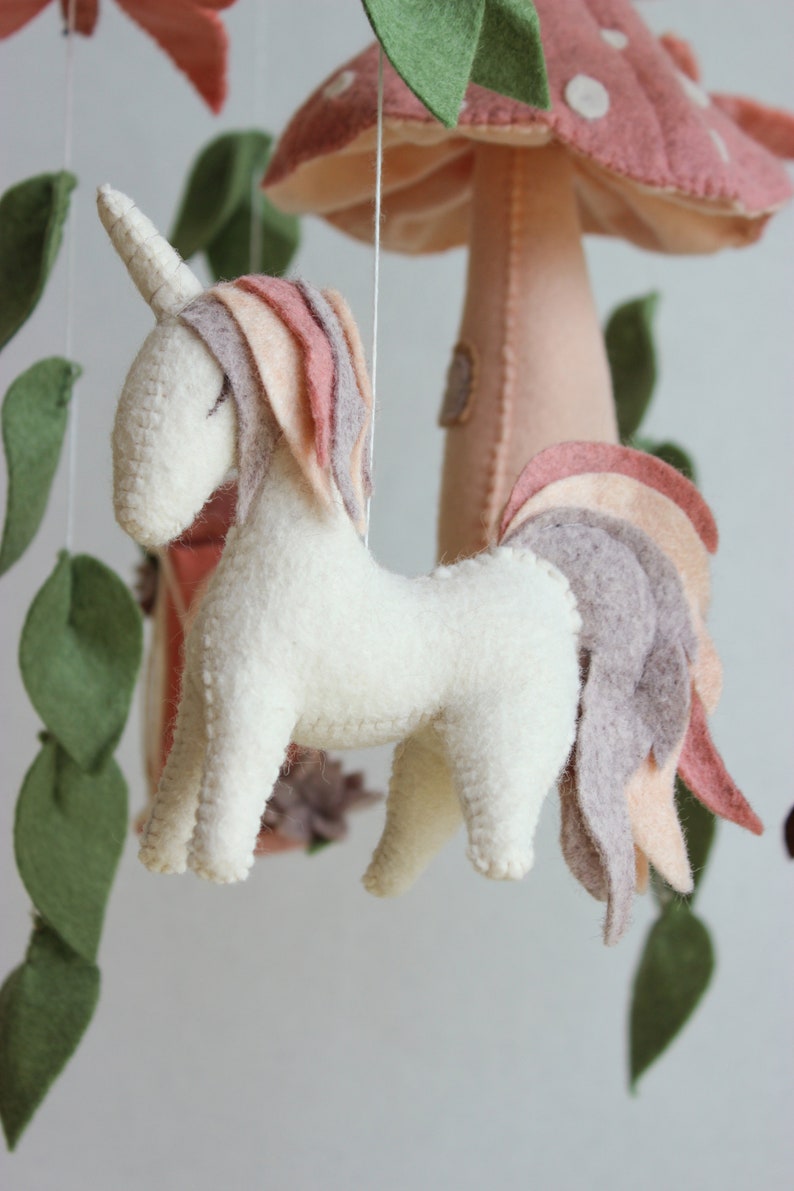 May include: A white felt unicorn with a pink and brown mane and tail hangs from a mobile. The unicorn is suspended from a white string. The mobile also features a pink felt mushroom with white spots and green felt leaves.