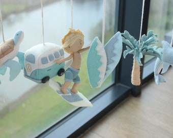 May include: A felt mobile with a surfer, a surfboard, a palm tree, a dolphin, a turtle, and a blue and white van. The mobile is hanging from a window.