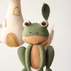 May include: A handmade felt mobile featuring a smiling green frog with a tan belly and large, round eyes. The mobile also includes a mushroom and leaf accents in shades of brown, cream, and green, creating a whimsical nursery decoration.