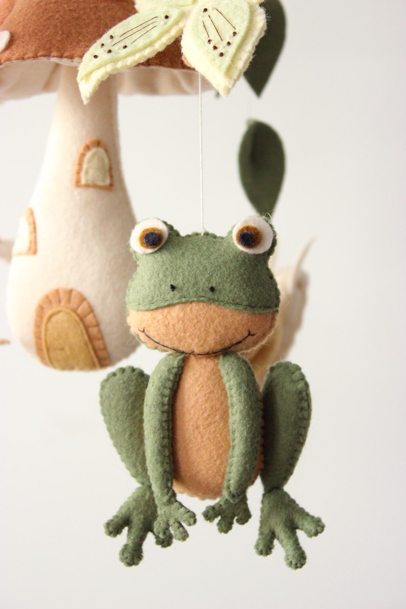 May include: A handmade felt mobile featuring a smiling green frog with a tan belly and large, round eyes. The mobile also includes a mushroom and leaf accents in shades of brown, cream, and green, creating a whimsical nursery decoration.