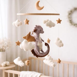 May include: A whimsical baby mobile featuring a felt dragon, clouds, stars, and a crescent moon. The dragon is lavender with brown wings and tail. The mobile is suspended from a wooden hoop, creating a dreamy nursery decoration.