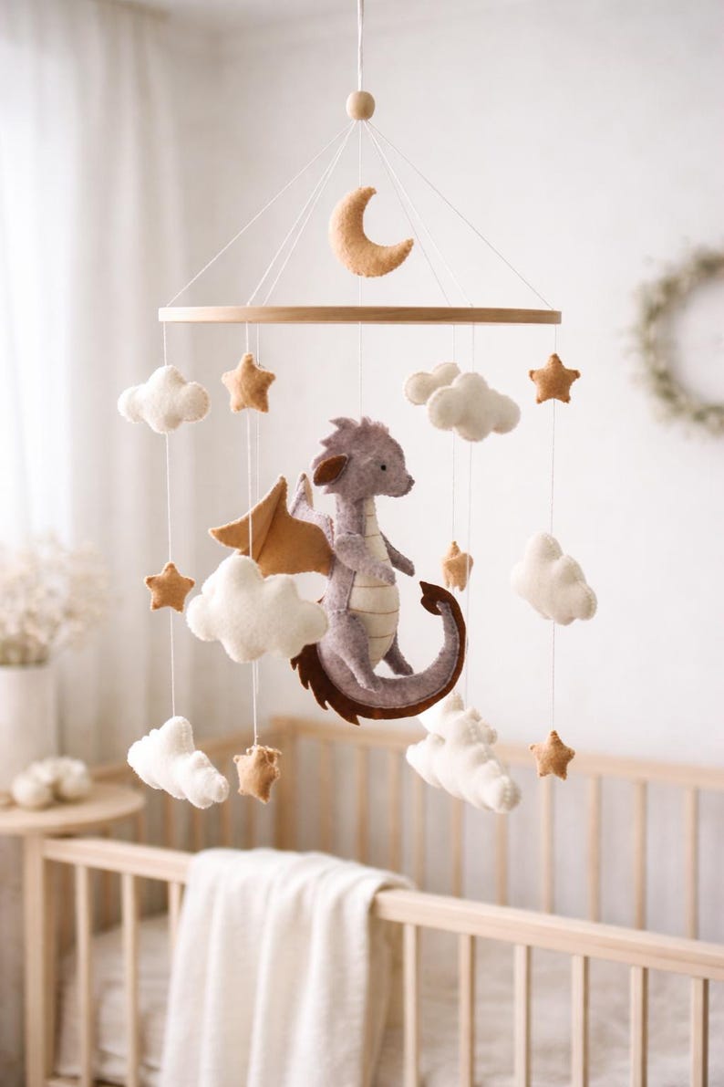May include: A whimsical baby mobile featuring a felt dragon, clouds, stars, and a crescent moon. The dragon is lavender with brown wings and tail. The mobile is suspended from a wooden hoop, creating a dreamy nursery decoration.