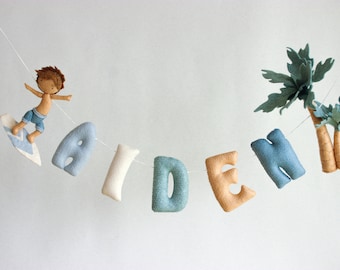 May include: A felt banner with the name "Aiden" spelled out in blue, white, and orange letters. A small felt surfer figurine is on the left side of the banner, and two palm trees are on the right side.