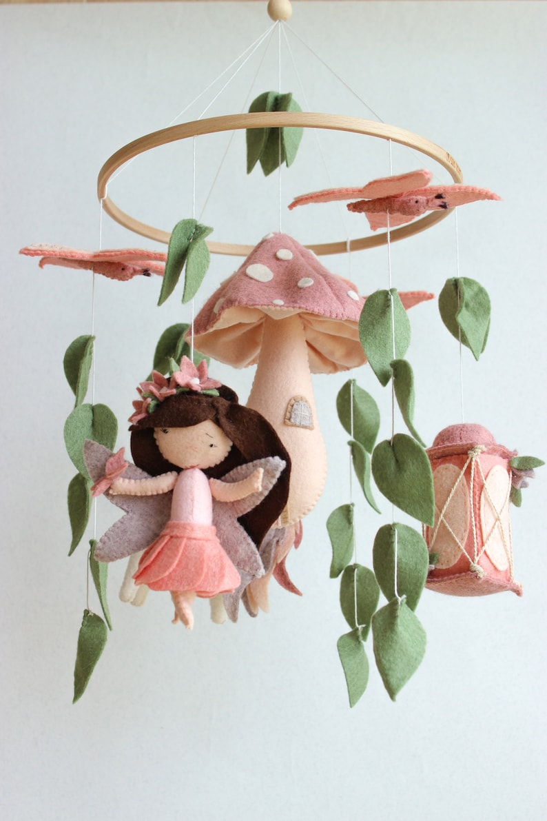 May include: A pink and green felt baby mobile with a fairy, mushroom, lantern, and leaves hanging from a wooden ring.