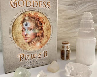 May include: A Goddess Power Oracle card deck with a 52-card deck and guidebook. The box features an illustration of a woman's face. Also shown are crystals, a small glass bottle, and a selenite tower.