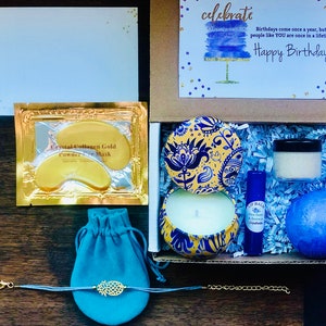 May include: A gift box with a birthday theme, containing a card with the words "Happy Birthday!", a gold eye mask, a blue velvet pouch, a bracelet, a candle, a lip balm, a jar of cream, and a blue bath bomb.