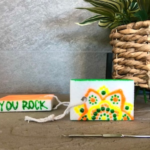 May include: Two white painted foam blocks with a string attached. One block has the words "YOU ROCK" written in green paint. The other block has a colorful mandala design with green, yellow, and orange paint.