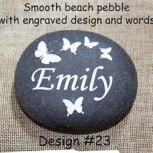 May include: A smooth gray beach pebble with a white engraved design of butterflies and the name "Emily". The pebble is on a beige burlap background. Design #23.