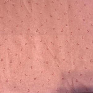 May include: A dusty rose fabric featuring a repeating pattern of small, dark, outlined triangles evenly spaced across the surface. The material has a soft, matte texture with visible folds and creases throughout the textile surface.