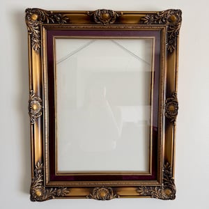May include: Ornate rectangular picture frame with a gold finish and burgundy inner border. The frame features detailed floral and scrollwork designs on the corners and edges. The frame is empty, with a clear glass pane.