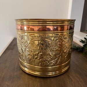 May include: A round, vintage brass planter with a copper band. The brass body features an embossed floral design. The planter is a decorative item for indoor use.