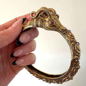 May include: Ornate gold-coloured ram head bracelet. The ram's head has a red eye and curled horns. The bracelet is decorated with blue accents and detailed patterns, giving it a vintage or antique appearance.