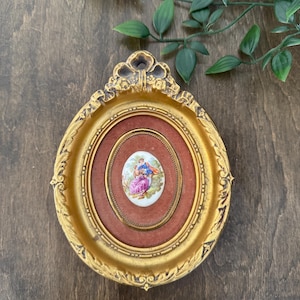 Puede incluir: Un marco ovalado dorado antiguo con una pequeña pintura detallada de una pareja en un jardín. La pintura está colocada sobre un fondo rojo dentro del marco. El marco tiene detalles ornamentados y se exhibe sobre una superficie de madera oscura.