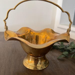 Peut inclure: Un panier en laiton vintage avec un bord festonné et une poignée incurvée. Le panier a une base de piédestal et une finition dorée brillante. La poignée est fixée aux côtés du panier avec de petites charnières. Le panier est vide.