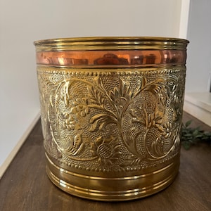 May include: A cylindrical brass container with intricate floral and leaf patterns. The top rim features a copper band, and the base and top are trimmed with a polished brass finish. The container is likely used for storage or decoration.