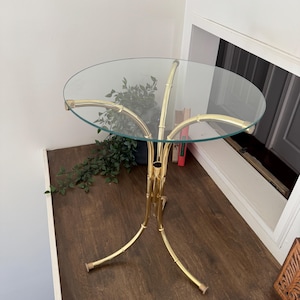 Puede incluir: Mesa auxiliar con tapa de cristal transparente y estructura metálica dorada. El marco tiene un diseño de estilo bambú. Las patas de la mesa se curvan hacia afuera. La mesa probablemente se utiliza con fines decorativos.