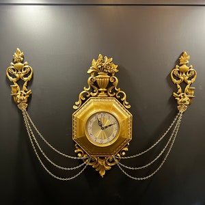 May include: Ornate gold-colored wall clock with an octagonal face and Roman numerals. The clock is flanked by decorative sconces and connected by chains. The clock has a classic, vintage design.