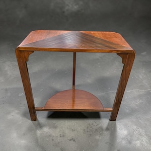 May include: A wooden corner table featuring a triangular top with a chevron wood grain pattern, two straight legs with carved diamond accents, and a lower semi-circular shelf supported by a single central leg, finished in a warm brown tone.